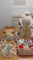 Wide view showing large Santa figure next to opened boxes containing vintage Christmas decorations such as paper angels and birds plus a box of gold beads.