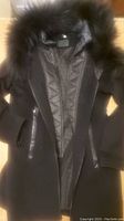 Full front view of black wool coat with fur-lined hood and visible double zipper and leather pocket trims.