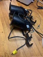 Two black metal industrial spotlights with clamps, safety wires, attached cables, and yellow 3-prong plug lying on a wooden floor.