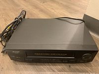 Front and top view of the Sharp 4-Head Hi-Fi Stereo VCR, black in color with visible power cord.