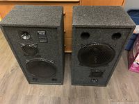 Pair of Audio Tech Pro Poly Series speakers shown front facing, covered in dark gray fabric, with visible speaker drivers and grills.