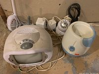 Photo showing two baby monitor receivers, one baby monitor camera unit, and one white NUK bottle warmer with blue accents. All units have power adaptors or cords visible.