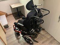 Rear view of the children's wheelchair showing the black cushioned seat, orange frame supports, large rear wheels, footrests, and push handles.