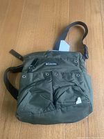 Front view of olive green Columbia Sportswear baby bag featuring logo patch, two buttoned front pockets, and adjustable black shoulder strap.