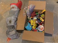 Photo showing the gray hanging IKEA fabric toy sorter with multiple circular compartments and red tips, next to a cardboard box filled with various toys including vehicles and figures.
