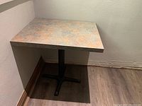 Square table with stone-effect laminate top, black metal pedestal base in a corner, showing the tabletop and base