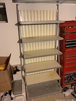 Full view of the utility shelf, showing the metal frame, six metal shelves, and the weighted pedestal base. Shelves appear disassembled or loose toward the bottom.
