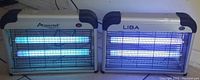 Pair of indoor electric insect killers with UV tubes turned on, one Aspectek branded and one Liba branded, plugged in and emitting blue light.