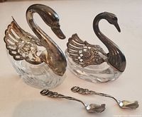 Front view of two swan-shaped salt and pepper cellars with glass bases and hinged silver necks and wings, along with two small sterling silver spoons placed in front.