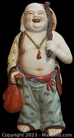 Front view showing the smiling Buddha holding a sack in the left hand and a walking stick over the right shoulder, colorful detailed painting on clothes.