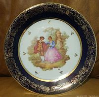 Front view of the round porcelain plate showing the deep cobalt border with ornate gold etchings and the central painted scene of the courting couple outdoors by a stone pillar.