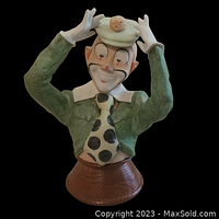 Front view of clown figurine wearing green jacket, polka dot tie, and small hat with pom-pom, mounted on wooden base.