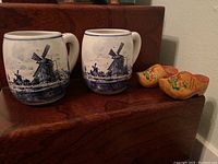Front view showing two Delft Blue ceramic mugs with windmill scenes and two small wooden clogs with floral paint.
