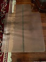 Translucent plastic floor protector mat placed on hardwood and rug floor, showing full size and shape with slight discoloration.