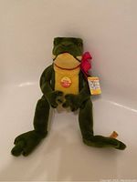 Front view of the green and yellow Steiff Cappy Frog plush showing the red bow and yellow tag on foot.