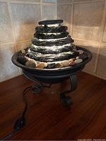 Side view showing the layered black stone waterfall, surrounding multicolored decorative stones, black basin, and scroll metal stand base with power cord and switch visible.