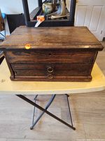 Closed view of rectangular antique wooden box with two front drawers and a hinged top lid, showing wood grain and condition issues.