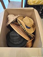 Top-down view of a cardboard box filled with various hats including a beige fedora, woven straw hats, a black felt hat, and a decorative sombrero.