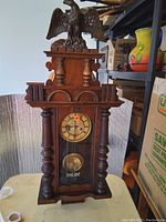 Full frontal view of the antique wooden wall clock on a white surface with surrounding shelving and boxes.