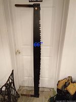 Full length image of the cross cut saw standing vertically, showing the overall length and shape with wooden handles and large teeth.