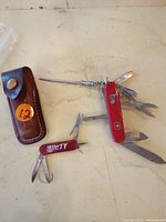 Photo showing vintage Swiss Army knife with various tools extended, smaller red multi-tool, and brown leather case.