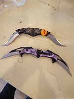 Two folding knives placed side by side on a surface; top knife with black handle and web design, bottom knife with purple handle and lightning design.