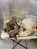 Overview of assorted items on a folding table including a wooden frame, decorative ceramic bowl, cables, packaging, and tools.