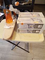 Four boxes of Hampton Bay mini pendant lights on a folding table with one unpacked Murano glass pendant light beside them.