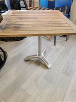 Full view of square teak patio table with aluminum base and teak inserts in base feet, showing discoloration and wear on the wood top.