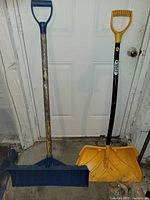 Photo showing two snow shovels: blue wooden handle shovel and yellow plastic ERA brand shovel