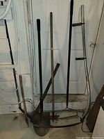 Full view of garden tools leaning against a white door, showing variety of shovels, rakes, and hoes with wooden handles and metal heads.