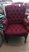 Front view of a burgundy upholstered wingback chair with tufted back and seat, brass nailhead trim on armrests, and cabriole wooden legs.