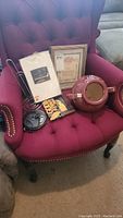 Items on a burgundy tufted armchair including a large red ceramic pot with lid, a framed religious print, disc player remote, and packing of greeting cards.