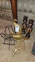 Lot with pair of golden peacock wall hangings, two wooden candle holders, two metal picture easels, and ornate metal wall sconce, arranged on carpet in front of couch.