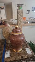 Photo showing brown crackled ginger jar with gold accents and tall beige carved vase in the background on a marble table.
