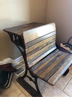 Front-side angled view of the antique wooden school desk with attached seat placed against a wall on tiled floor.
