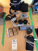 Overview photo showing Bushnell binoculars with strap and carrying case, black and silver portable speaker, two Coors Light power banks, small stereo speaker keychain, black round lenses, small packaged camera, and assorted cables.