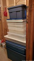Stack of six plastic storage bins of varying colors and sizes, stacked five deep against an unfinished wood wall with basement surroundings.