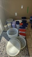 Overview of 20 plastic food storage containers of various sizes and shapes placed on floor and rug near wall.