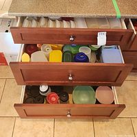 Three kitchen drawers pulled open showing a variety of plastic food storage containers, water bottles, and travel mugs.
