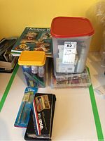 Overview of polymer clay modeling books and crafting supplies in plastic storage containers with yellow and red lids, plus an X-Acto knife and blister pack of blades.