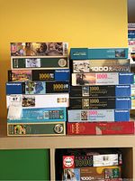 Stacked puzzle boxes showing mostly 1000-piece puzzles from Ravensburger, Eurographics, Educa, Trefl and other brands. Themes range from animals, space, village scenes, cities and historical images.