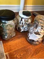 Three glass jars on wooden table filled with assorted shells and rocks. One jar has a dark rock on the lid.
