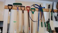 Wide view of multiple long-handled garden tools including shovels, broom, rake, loppers, and trimmer edger hung on wall