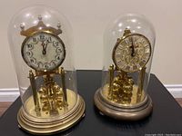 Front view showing two torsion clocks under glass domes on a dark flat surface.
