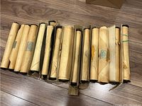 14 assorted player piano music rolls lying side by side on a wooden floor. Rolls are wrapped in aged paper with some labels and markings.