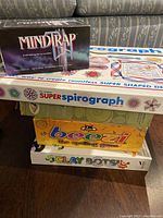 Stacked boxes of five games and toys showing titles MindTrap II, Super Spirograph, Beezi, Clay Bots