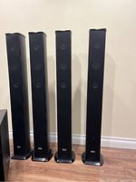 Four tall black speakers standing upright on hardwood floor against white wall