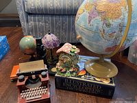 Photo showing large and small vintage globes, a purple umbrella lamp, mushroom music box on top of illustrated encyclopedia dictionary, and miniature red typewriter.