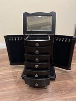 Front view of the black wooden jewelry box with top glass lid open, two side doors open revealing hooks, and six open drawers showing interior compartments.